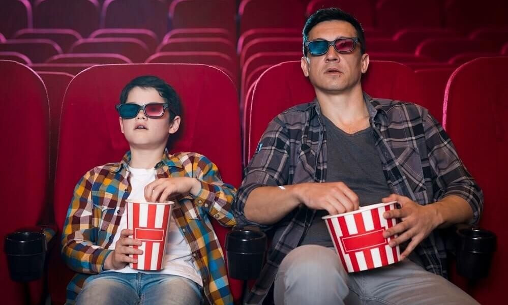 A boy and a young man are watching a Latin film in the cinema while eating popcorn.